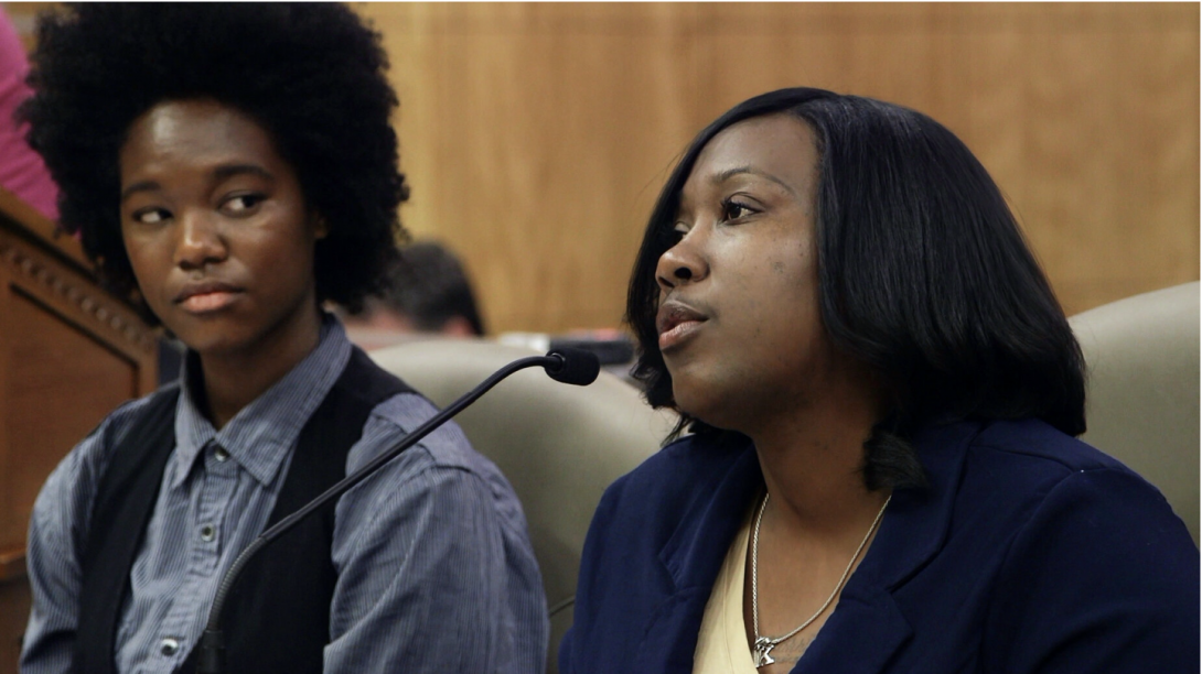Two black women sitting. One is speaking into a microphone