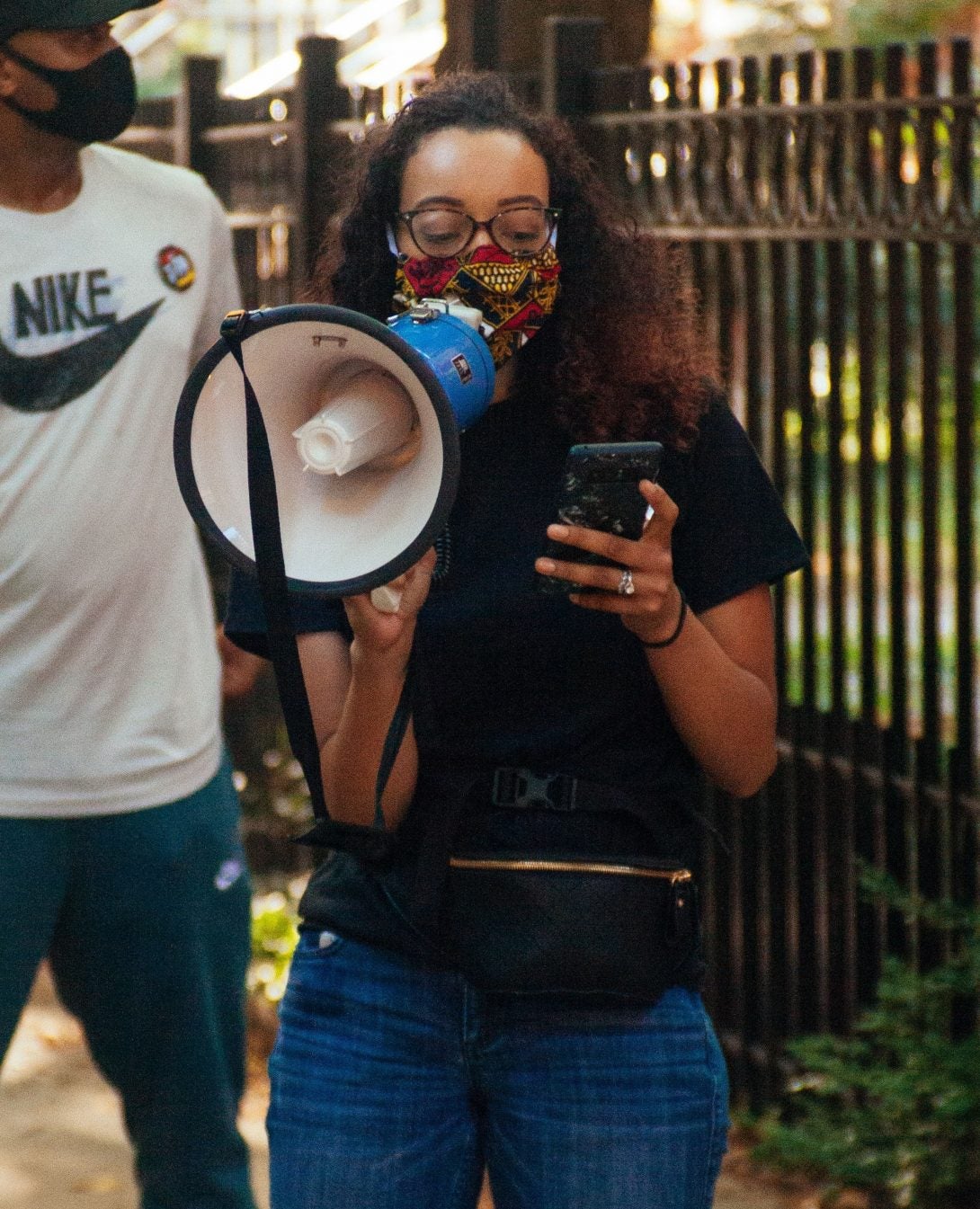 Alexis, wearing a face mask, standing outdoors near a fence, reading off a phone, and speaking through a megaphone.