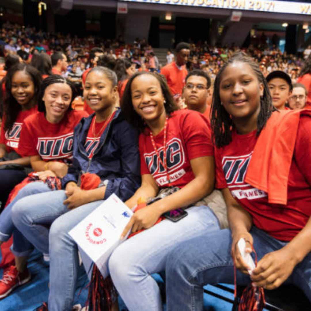 A group of UIC students in red UIC shirts sitting together at a large campus event and smiling toward the camera.