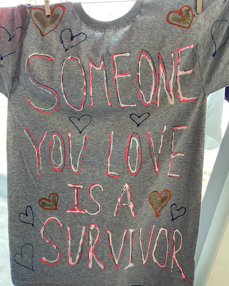 Pink and white painted text on a gray t-shirt reads, “Someone you love is a survivor” in all caps, surrounded by blue, pink, and gold hearts.