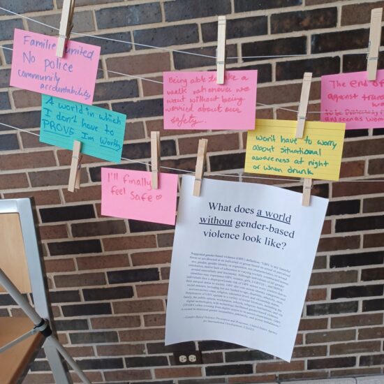 Written note cards hang from a clothesline as part of the community writing activity Omar led at the Survivors Belong Here Clothesline Project exhibit. Also hanging from the clothesline is a sheet with the activity title: “What does a world without gender-based violence look like?”