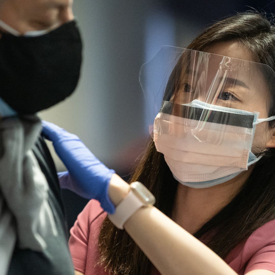 A person wearing a mask and face shield vaccinating another person wearing a mask.